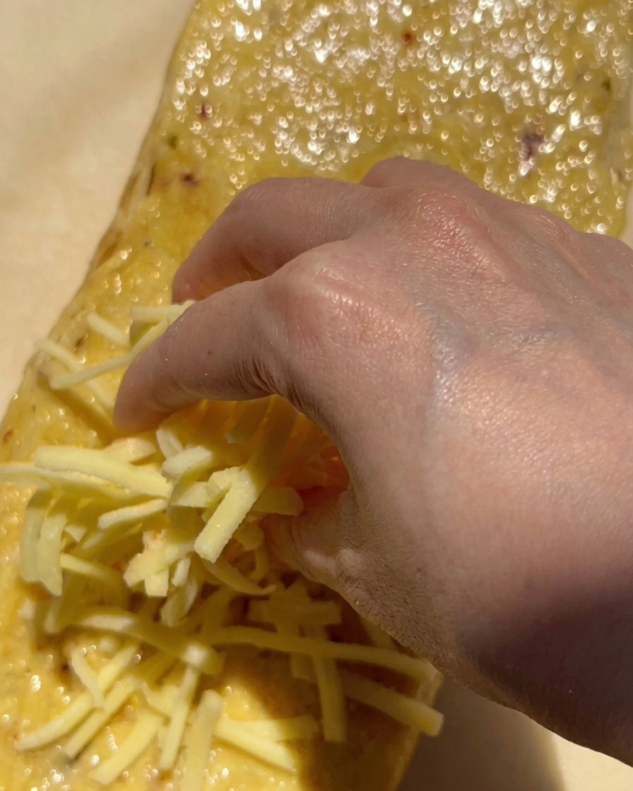A hand sprinkling a large handful of shredded white mozzarella cheese onto a loaf of bread spread with garlic butter.