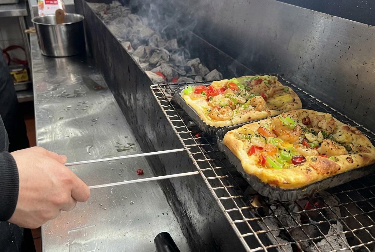 A rectangular pizza topped with shrimp and mixed vegetables being placed on a long charcoal grill grate inside a professional kitchen.