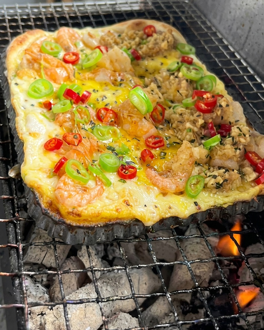 A rectangular pizza crust topped with shrimp and vegetables cooking over glowing charcoal embers.
