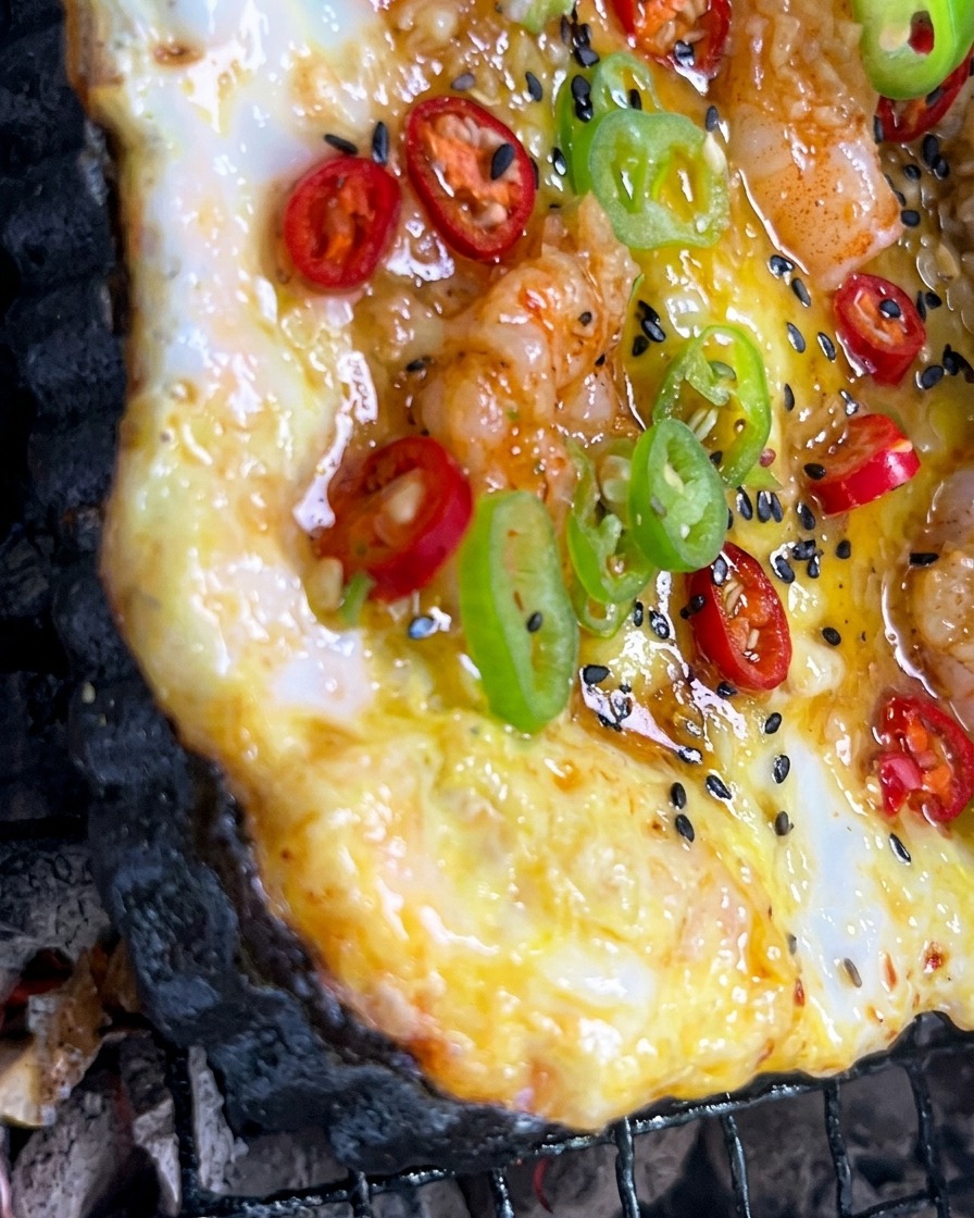 Minced garlic sauce being brushed onto a colorful pizza cooking over a charcoal fire.