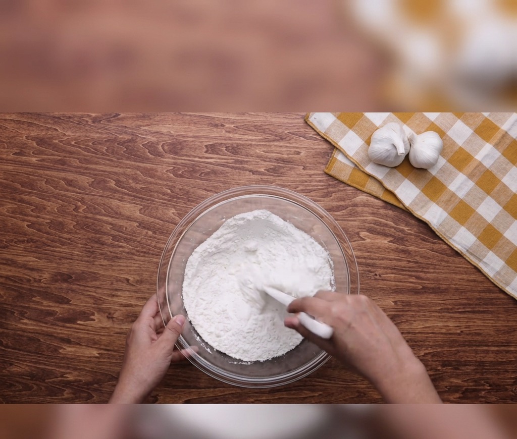Hands using a white spatula to mix flour, water, and oil in a glass bowl placed on a wooden tabletop.