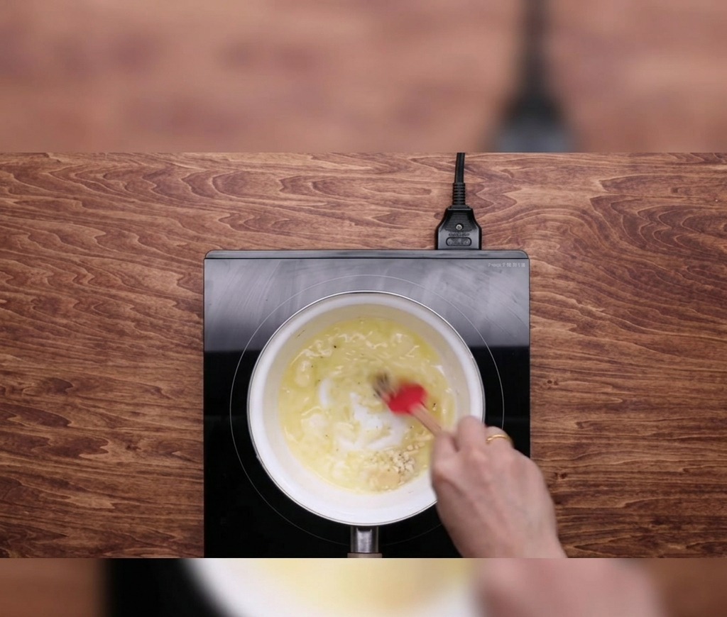 A hand using a red spatula to stir melted butter and minced garlic in a small white saucepan on a black induction cooktop.