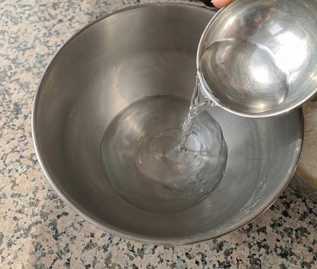Water being poured from a small metal bowl into a large stainless steel mixing bowl on a granite countertop.