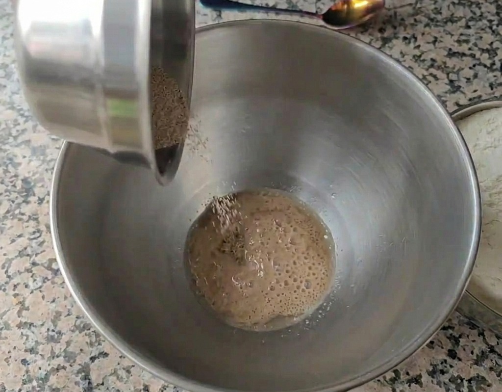 Dry granulated yeast being poured into a stainless steel bowl filled with water showing small bubbles forming on the surface.
