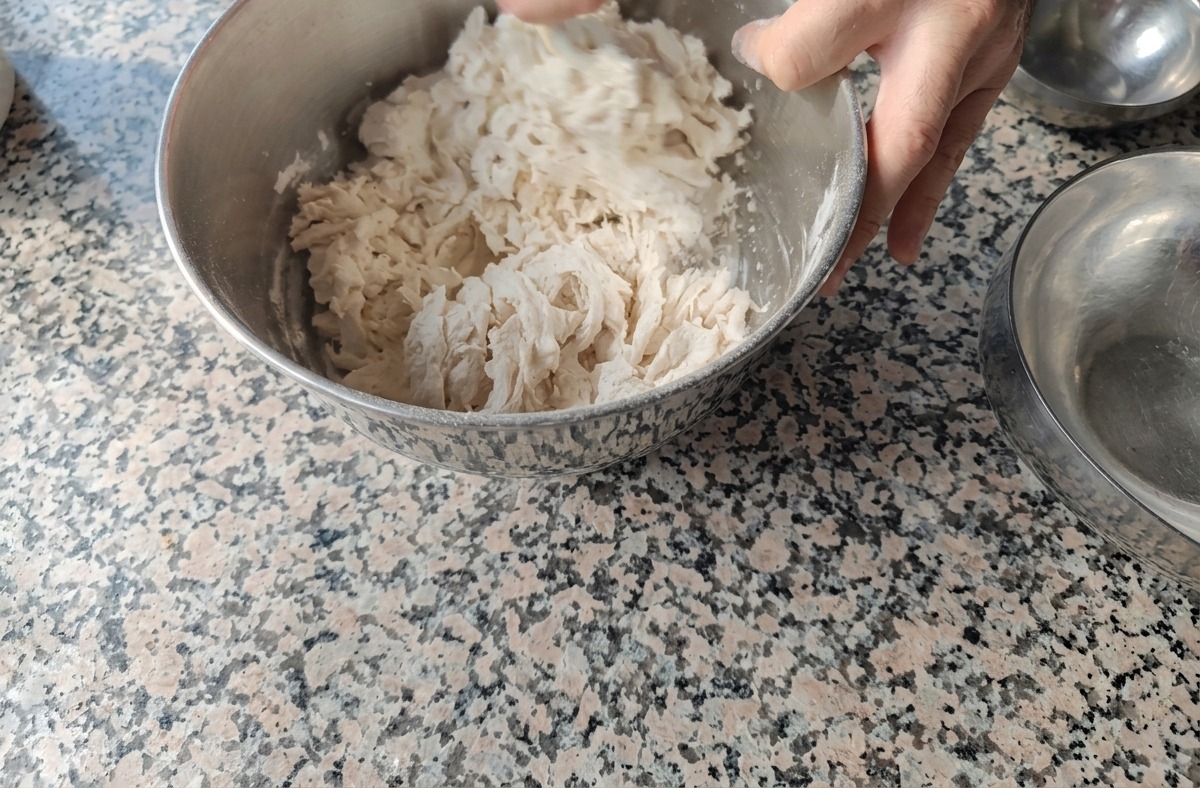 Hands mixing flour and water in a metal bowl to create a rough and shaggy Biga dough.