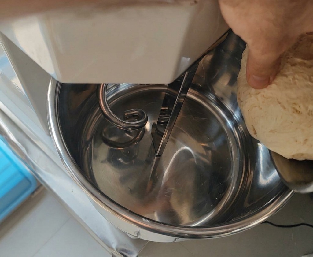 A hand placing a portion of fermented, textured Biga dough into a large industrial-style mixer bowl.