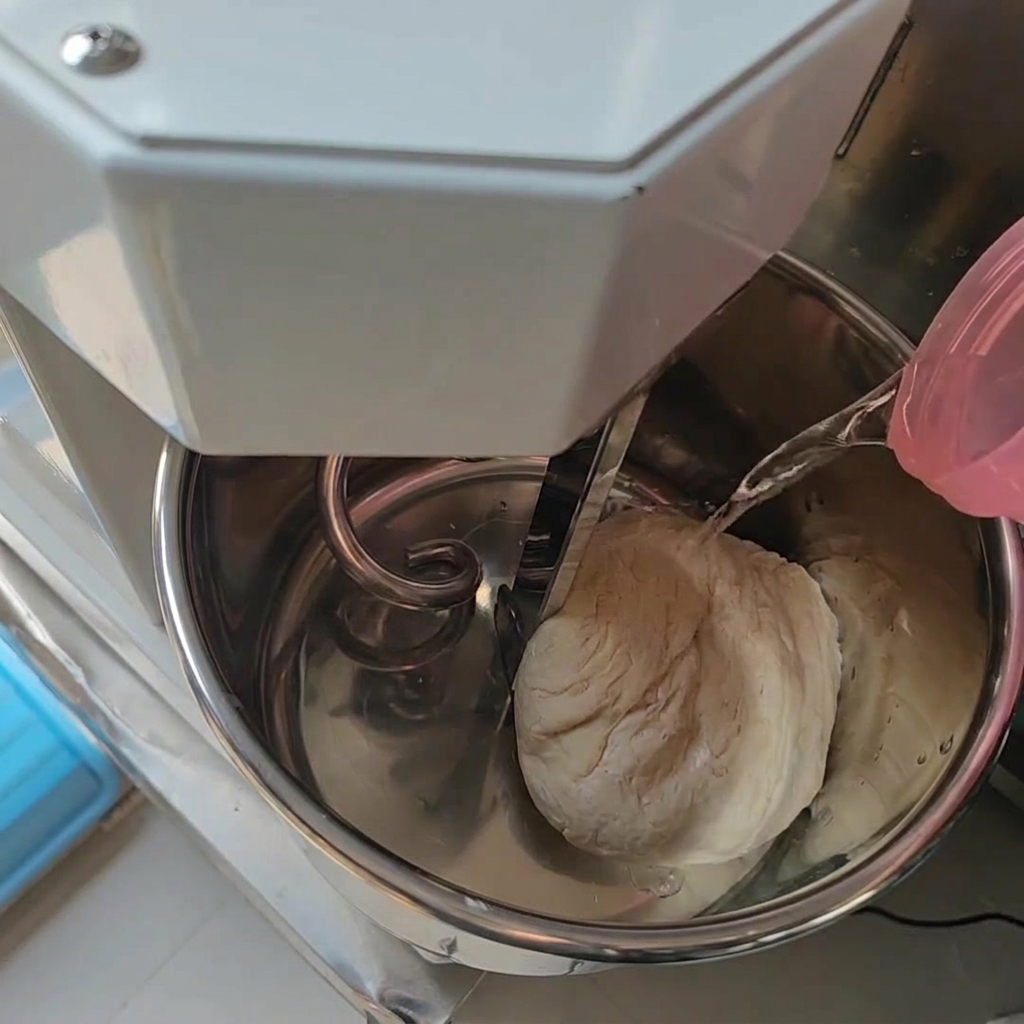 Water being poured from a pink container into a stainless steel mixer bowl that already contains a ball of dough.