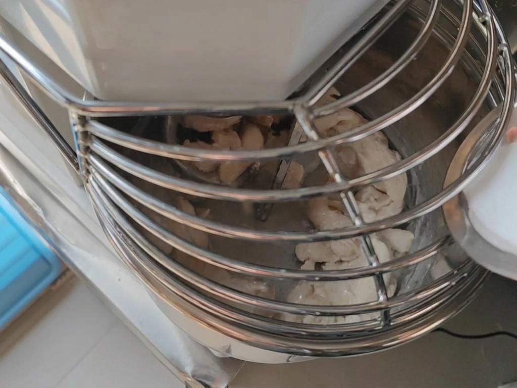 A small container pouring fine salt into a spiral mixer bowl where pizza dough is being kneaded.