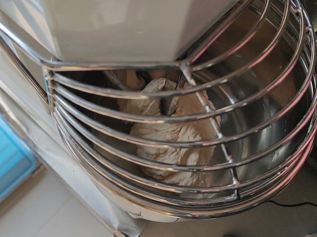 A thin stream of water being poured into a spinning mixer bowl containing developing pizza dough.