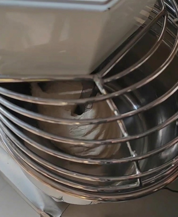 A professional spiral mixer containing a large smooth batch of pizza dough under a metal safety guard.