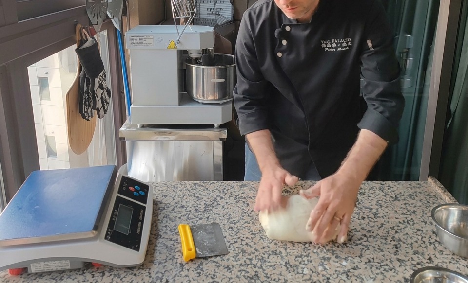 A large mass of pale pizza dough being divided with a yellow bench scraper on a speckled granite countertop next to a digital scale.