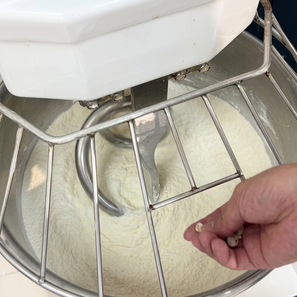 Hand crumbling pieces of fresh yeast into the flour inside a running spiral mixer.