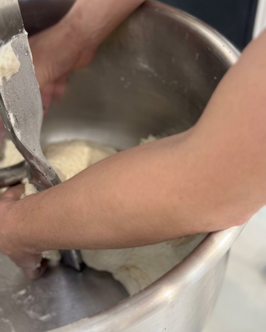 Hands pulling a large, cohesive mass of raw dough out of a metal spiral mixing bowl.