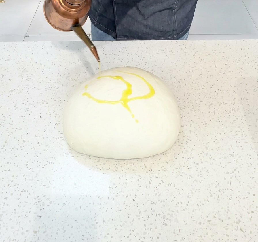 Chef drizzling olive oil from a copper pot onto a large, smooth ball of dough.