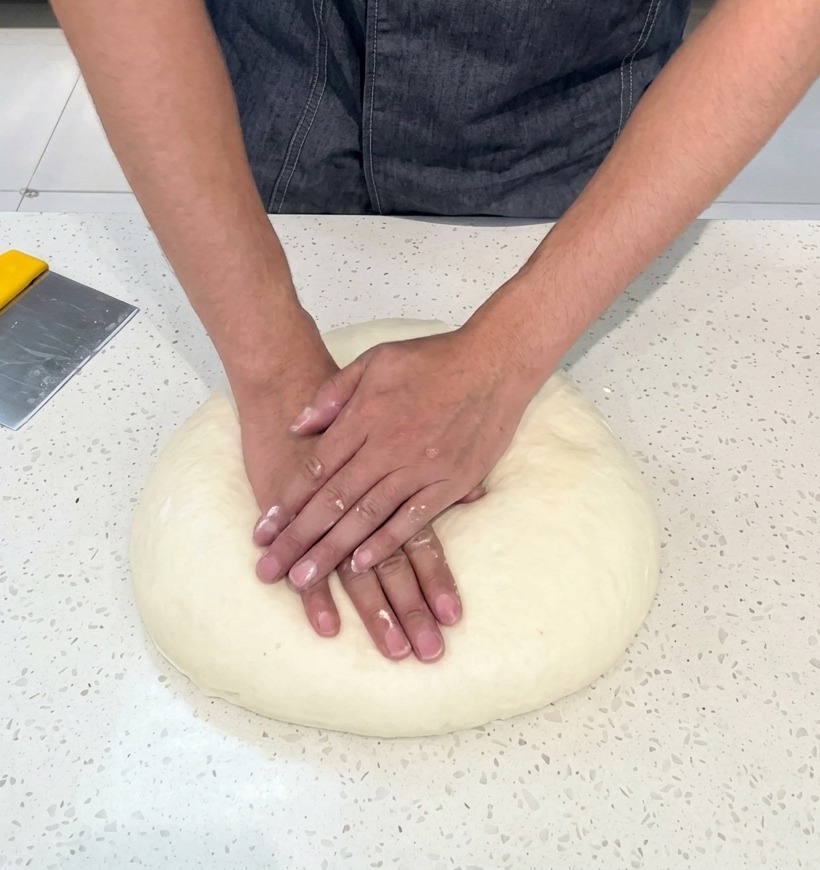 A person in a grey chefs coat using both hands to press down and flatten a large, smooth round of pale pizza dough on a white countertop.