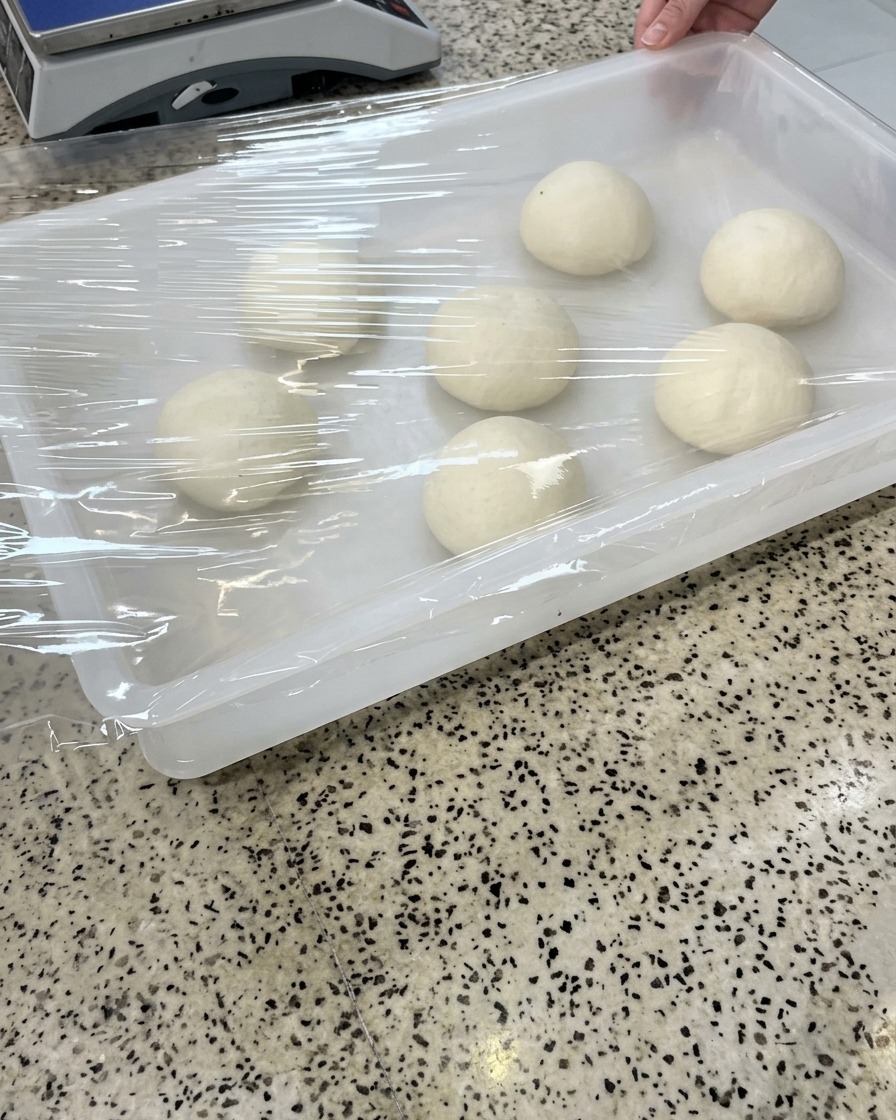 A white plastic tray containing six smooth dough balls being covered with a sheet of transparent plastic wrap.