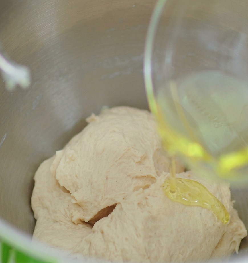 A stream of golden olive oil being poured from a small glass bowl onto a smooth ball of pizza dough.