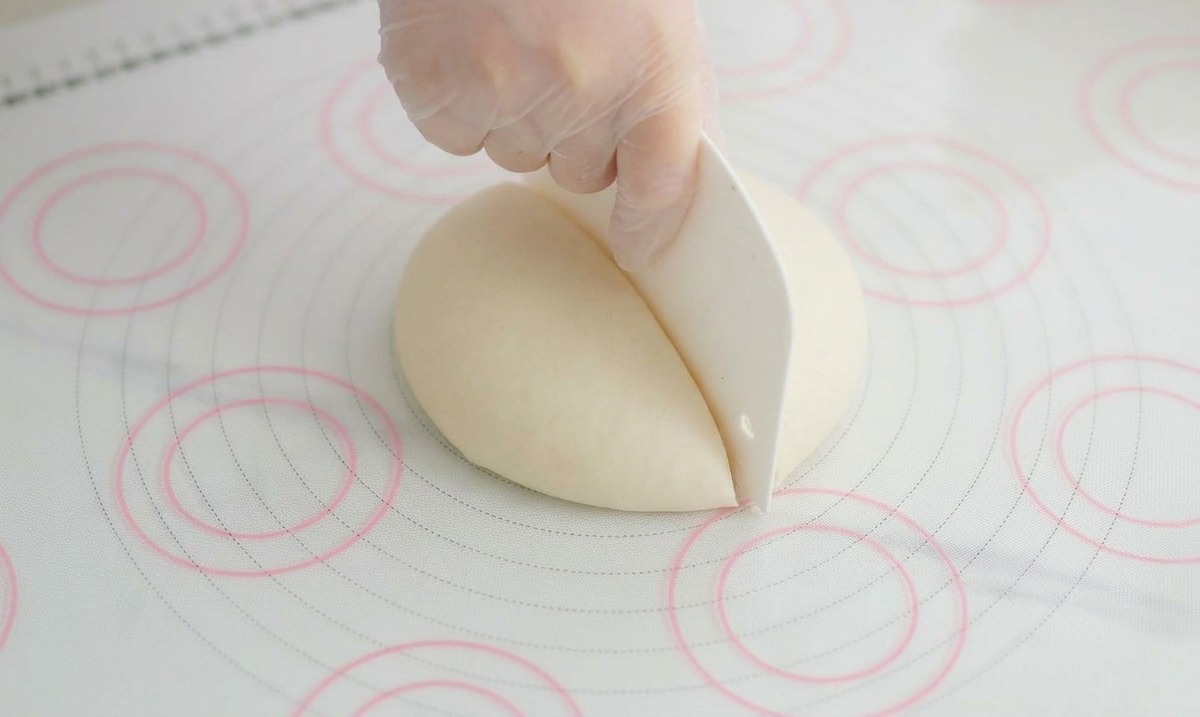 A hand using a white plastic bench scraper to divide a large smooth ball of pizza dough in half on a silicone mat.