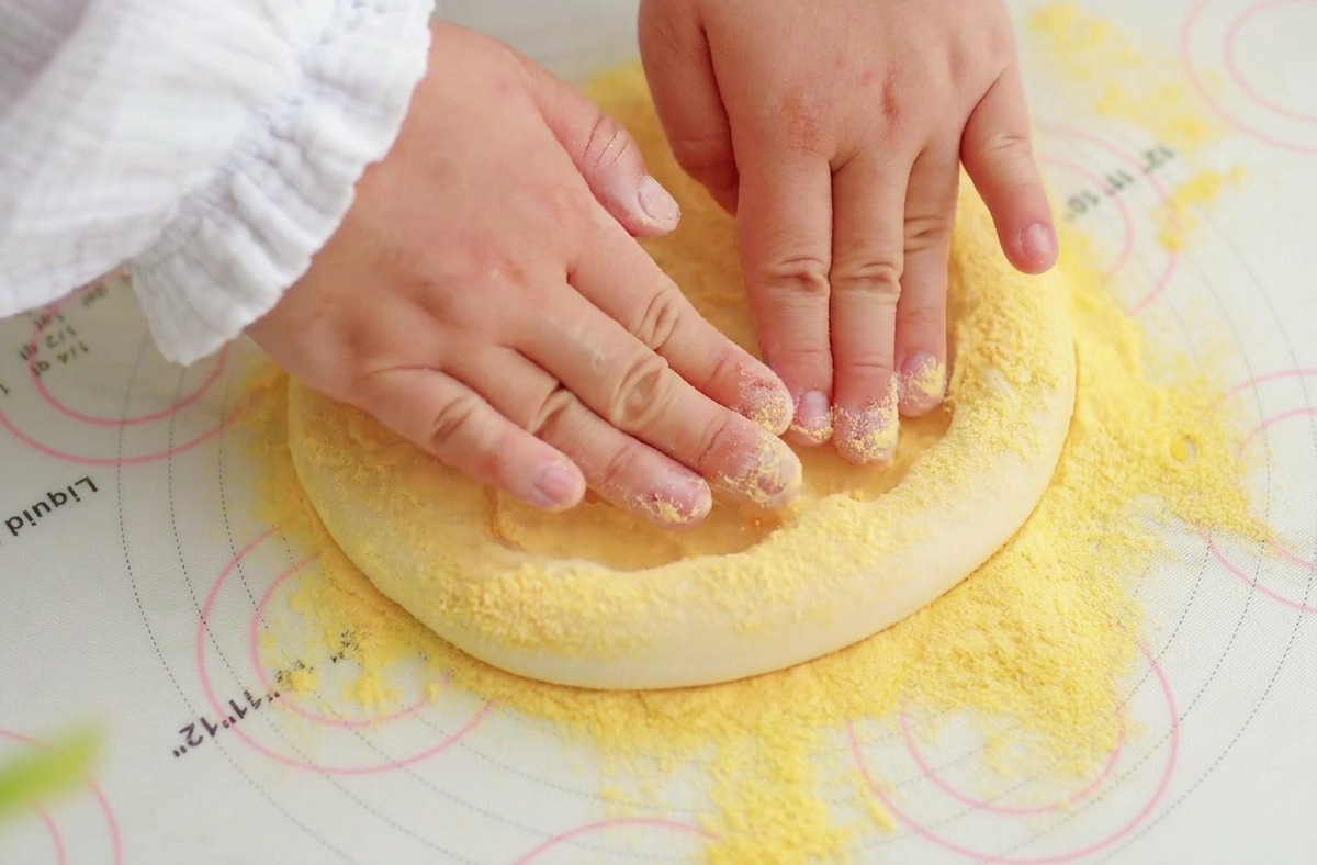 A pair of hands pressing the center of a pizza dough disc on a floured mat, pushing air toward the edges.
