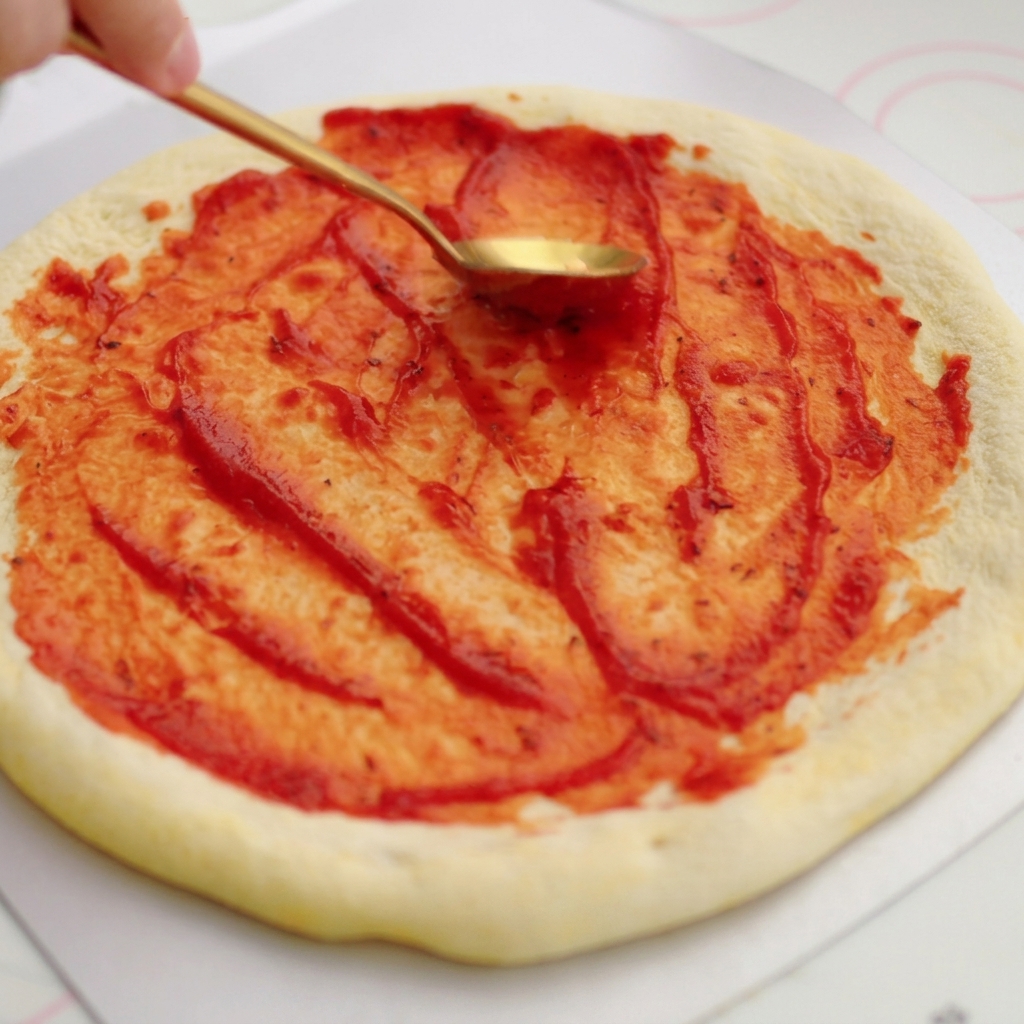 A golden spoon spreading bright red tomato sauce in circular motions onto a raw pizza base.