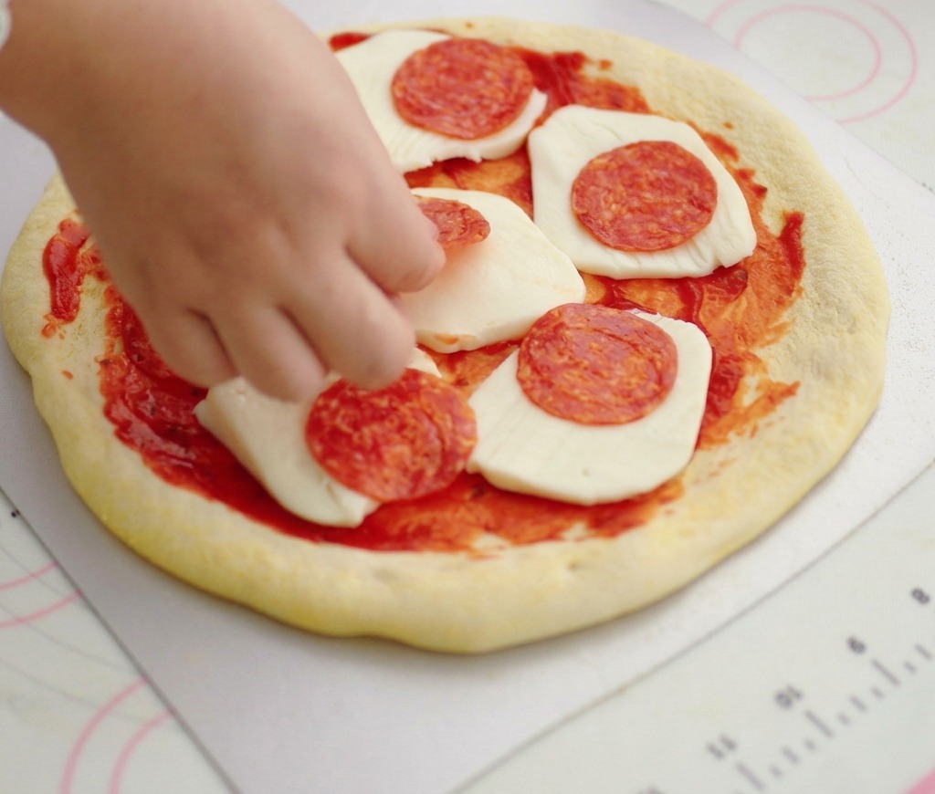 A hand placing thin round slices of salami onto a pizza crust topped with tomato sauce and fresh mozzarella cheese.