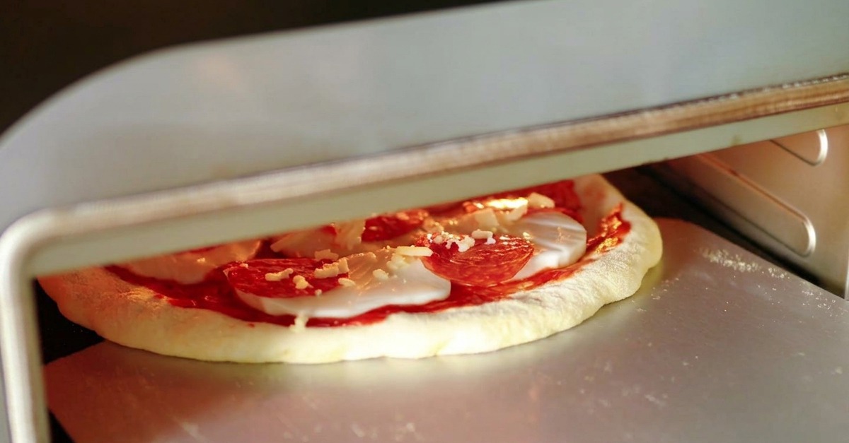 A raw salami pizza being transferred from a silver metal pizza peel into the glowing mouth of a high-temperature pizza oven.