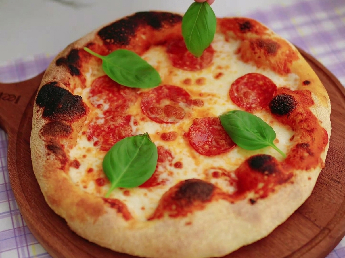 A hand placing fresh green basil leaves onto a freshly baked Neapolitan pizza featuring a charred crust and melted mozzarella.