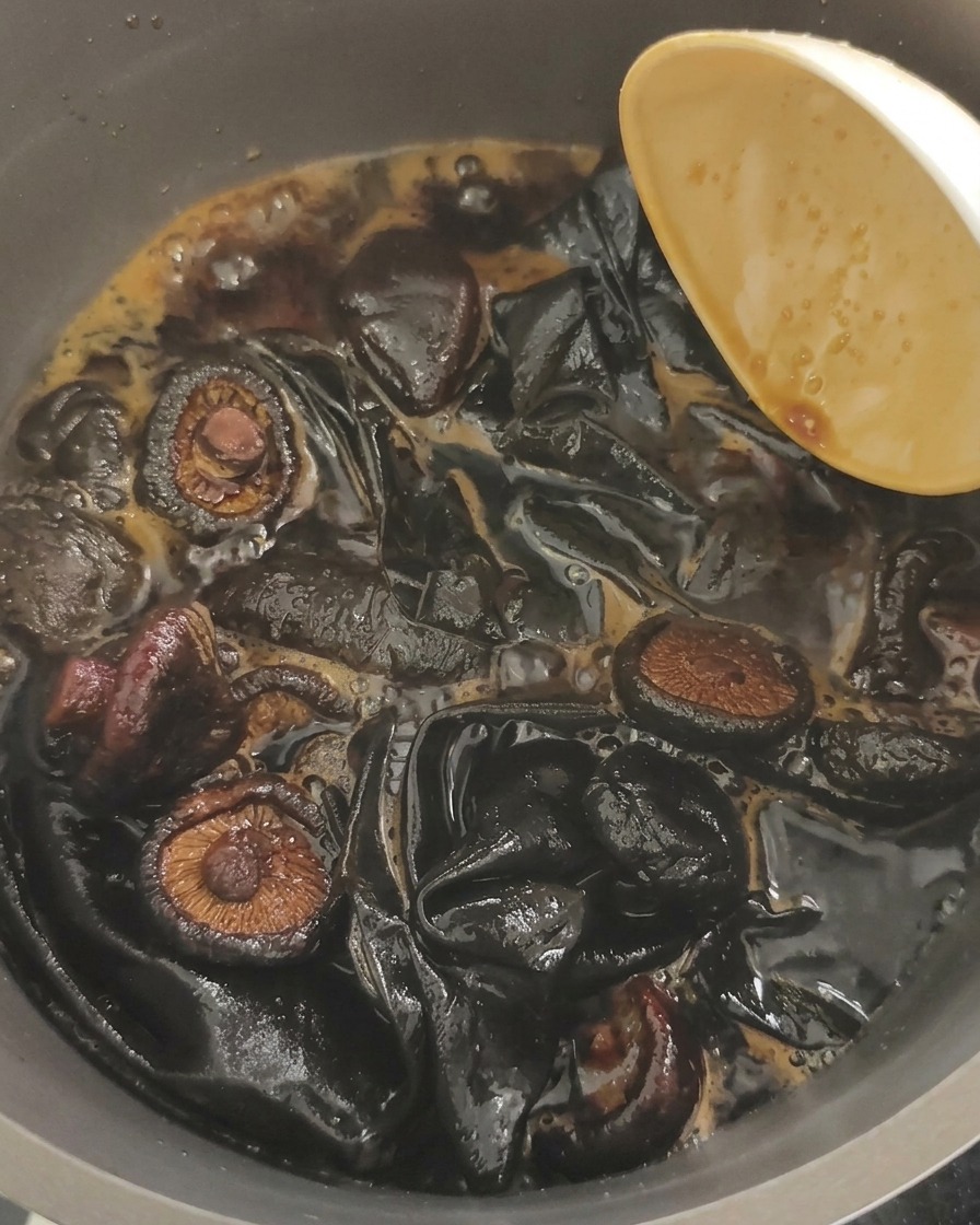 A close-up shot of wood ear mushrooms and large pieces of kombu simmering in a bubbling dark brown soy liquid.
