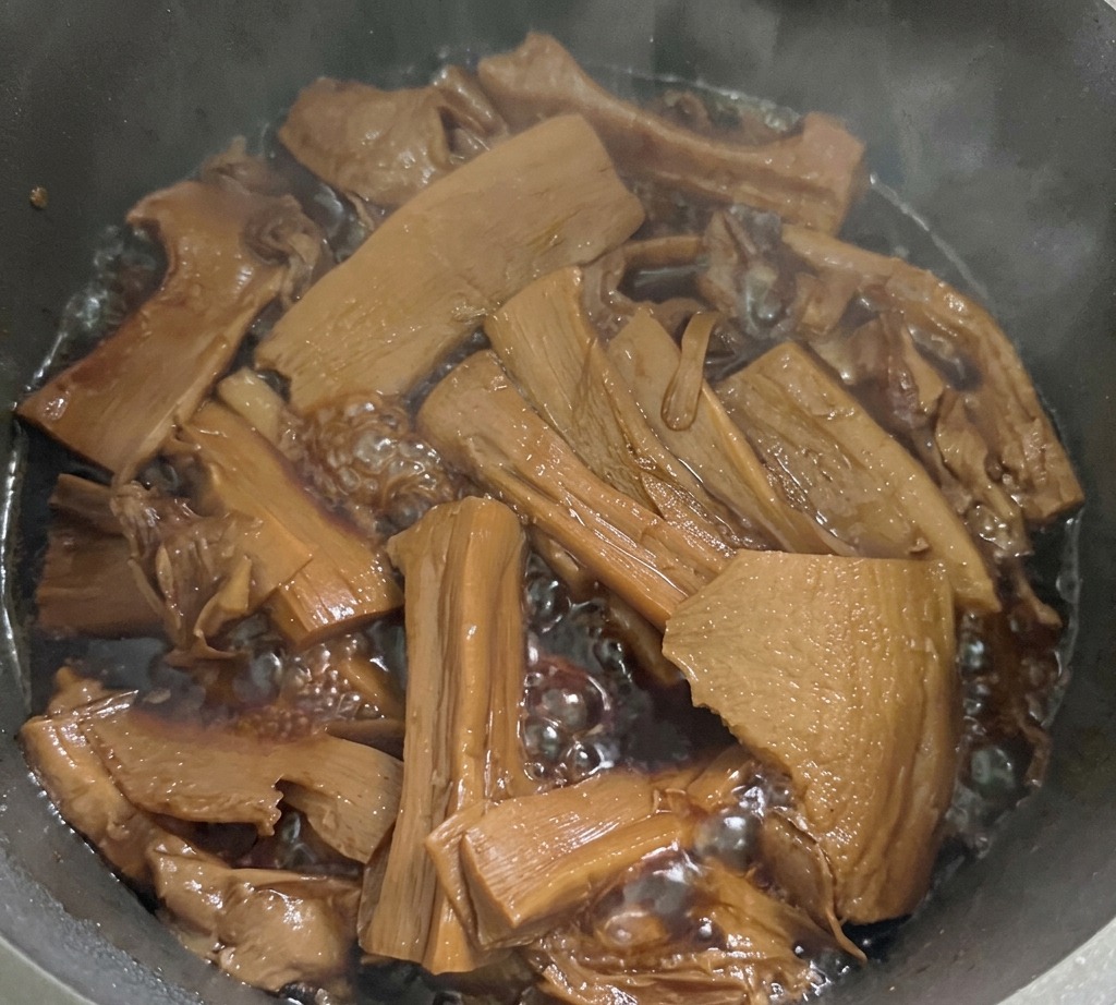 Sliced bamboo shoots simmering in a bubbling dark brown seasoning liquid inside a metal pot on the stove.