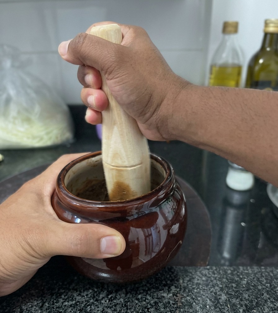 A person using a wooden pestle to grind dark, fried garlic inside a small brown ceramic jar on a granite countertop.