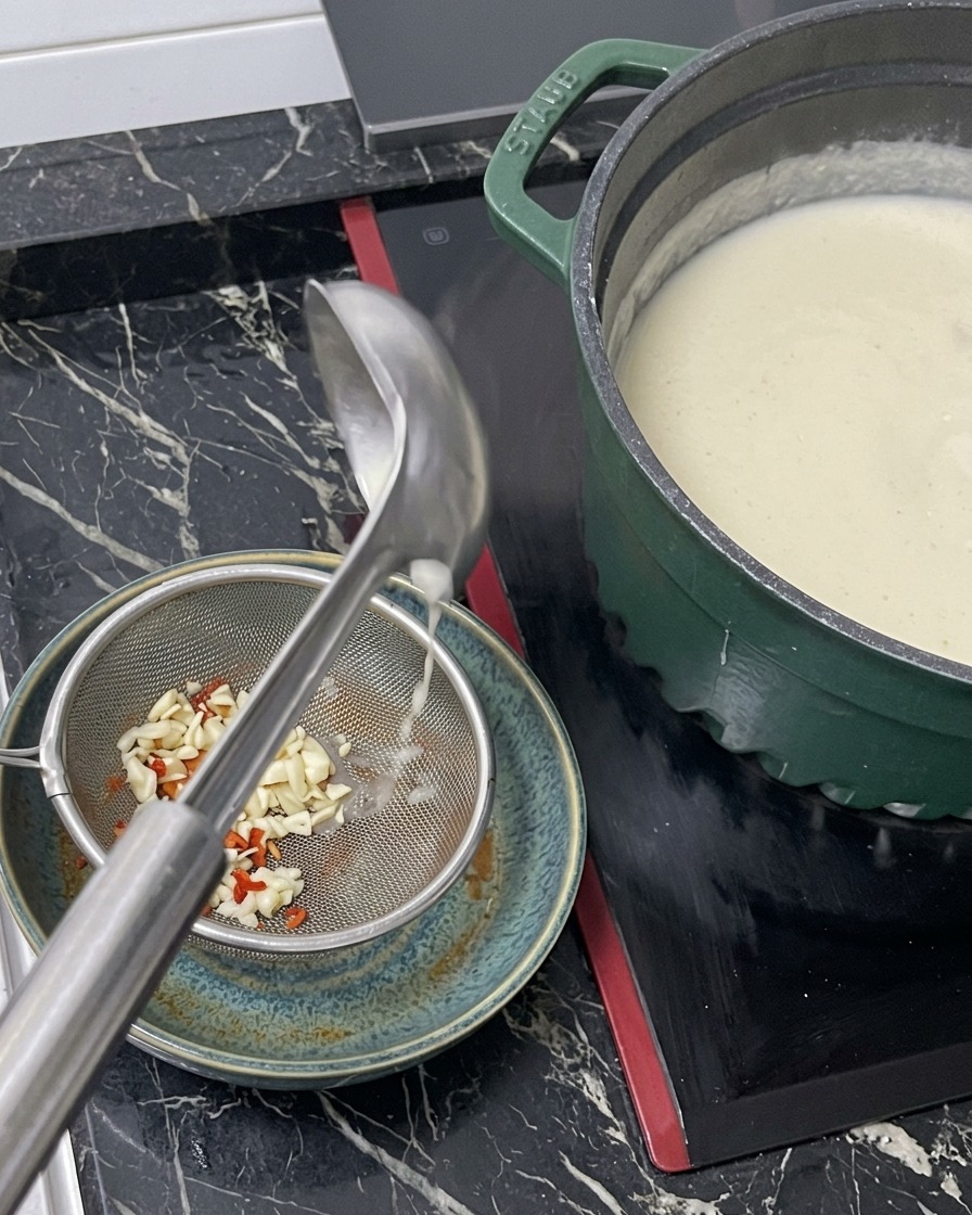 Hot white broth being poured from a ladle through a metal strainer into a bowl containing the ramen seasoning base.