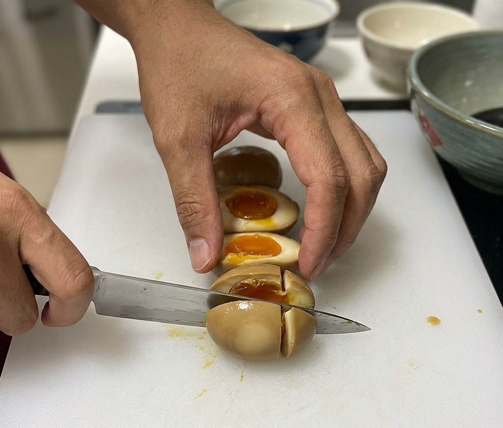 A chef slicing a brown, marinated soft-boiled egg in half on a white cutting board, showing the runny orange yolk inside.