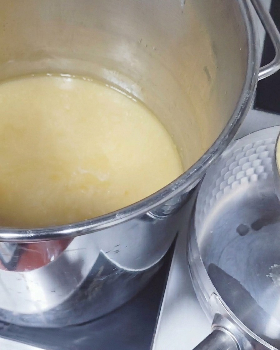 A large metal ladle lifting opaque, pale yellow pork broth from a deep stockpot and pouring it into a smaller saucepan.