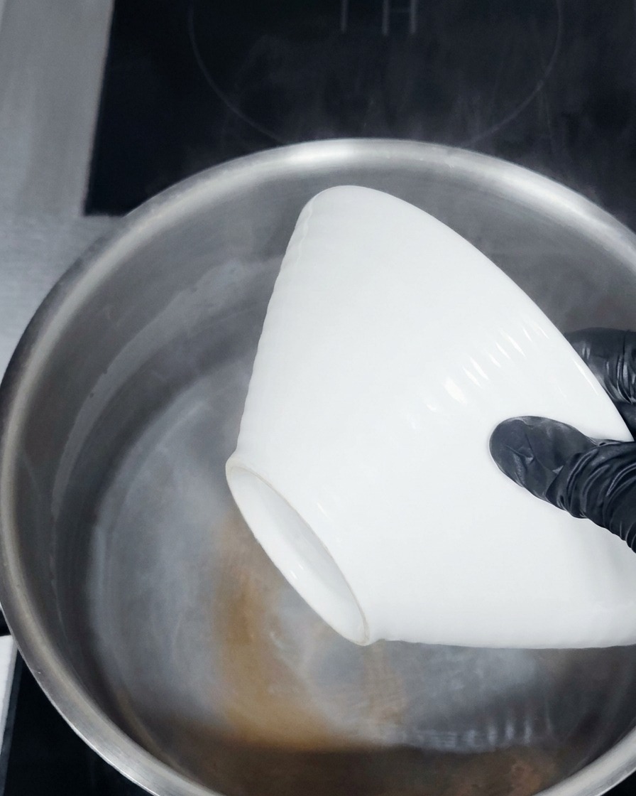Gloved hands lowering a white ceramic bowl into a pot of steaming hot water.