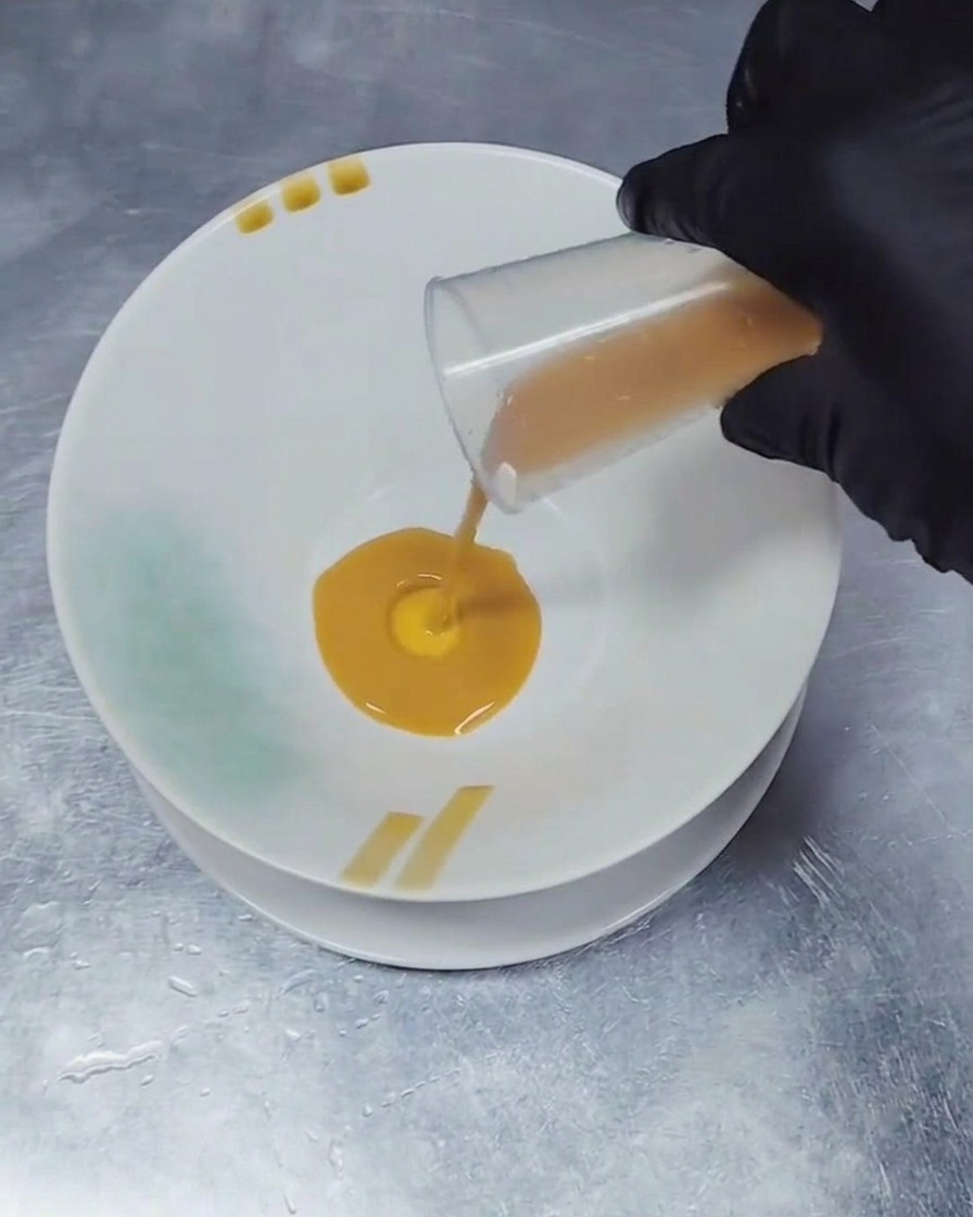 A gloved hand pouring a small amount of dark brown liquid seasoning from a clear measuring beaker into the center of a white ceramic bowl.
