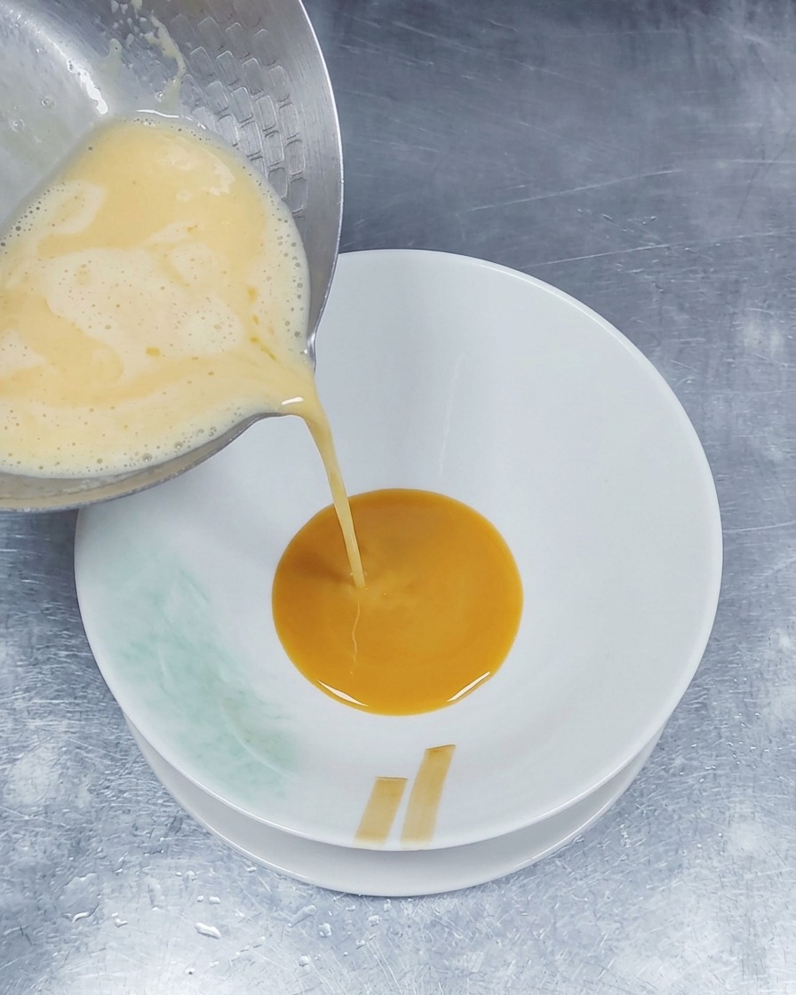 Hot, frothy pork bone broth being poured from a metal pot into a bowl that already contains a layer of brown seasoning sauce.