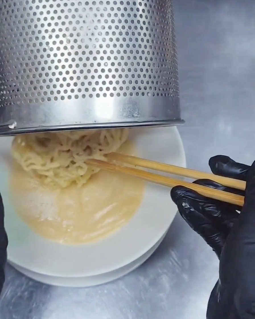 A stainless steel noodle strainer pouring wavy ramen noodles into a bowl of creamy Tonkotsu broth.