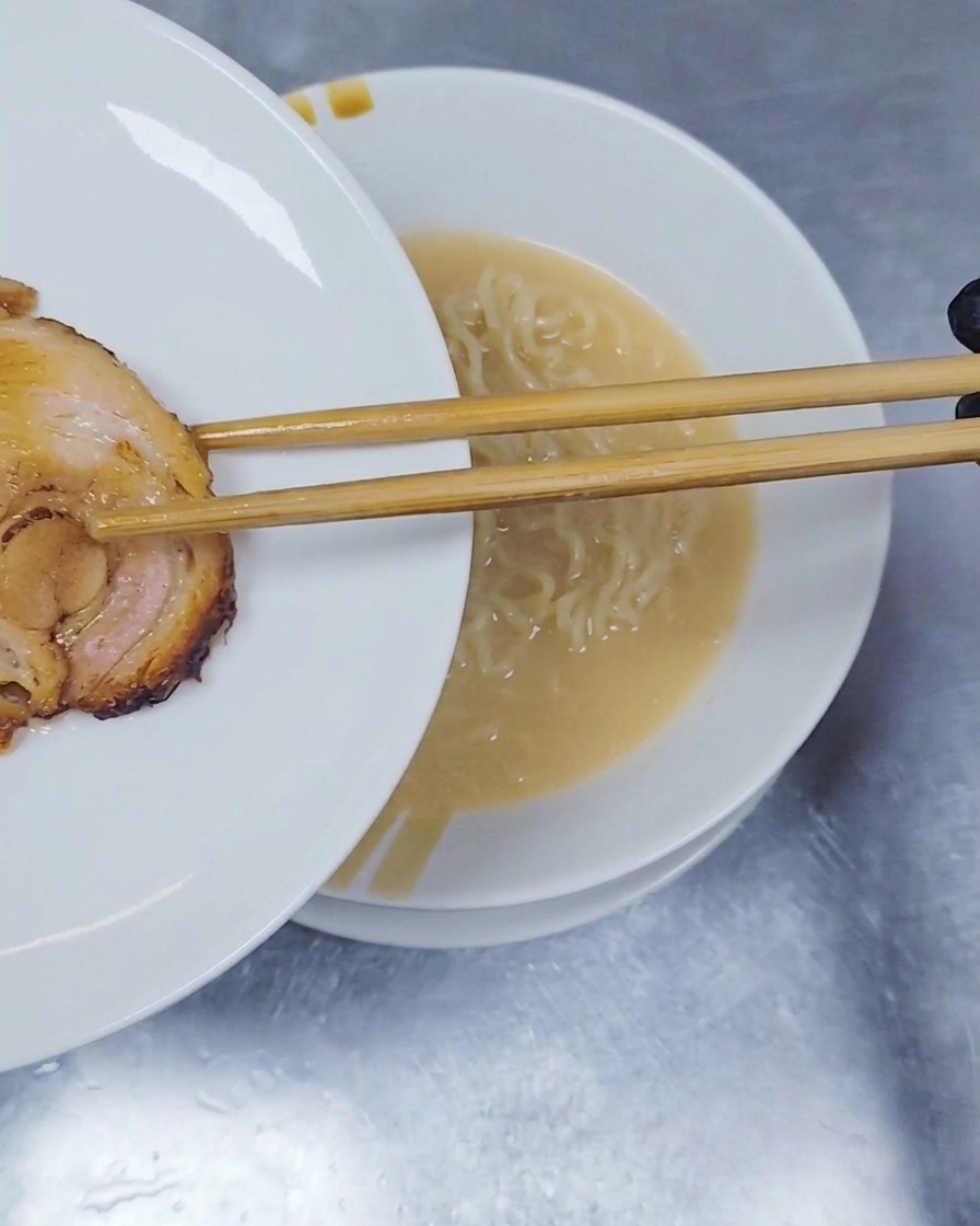 A slice of rolled and braised chashu pork being placed on top of ramen noodles with wooden chopsticks.