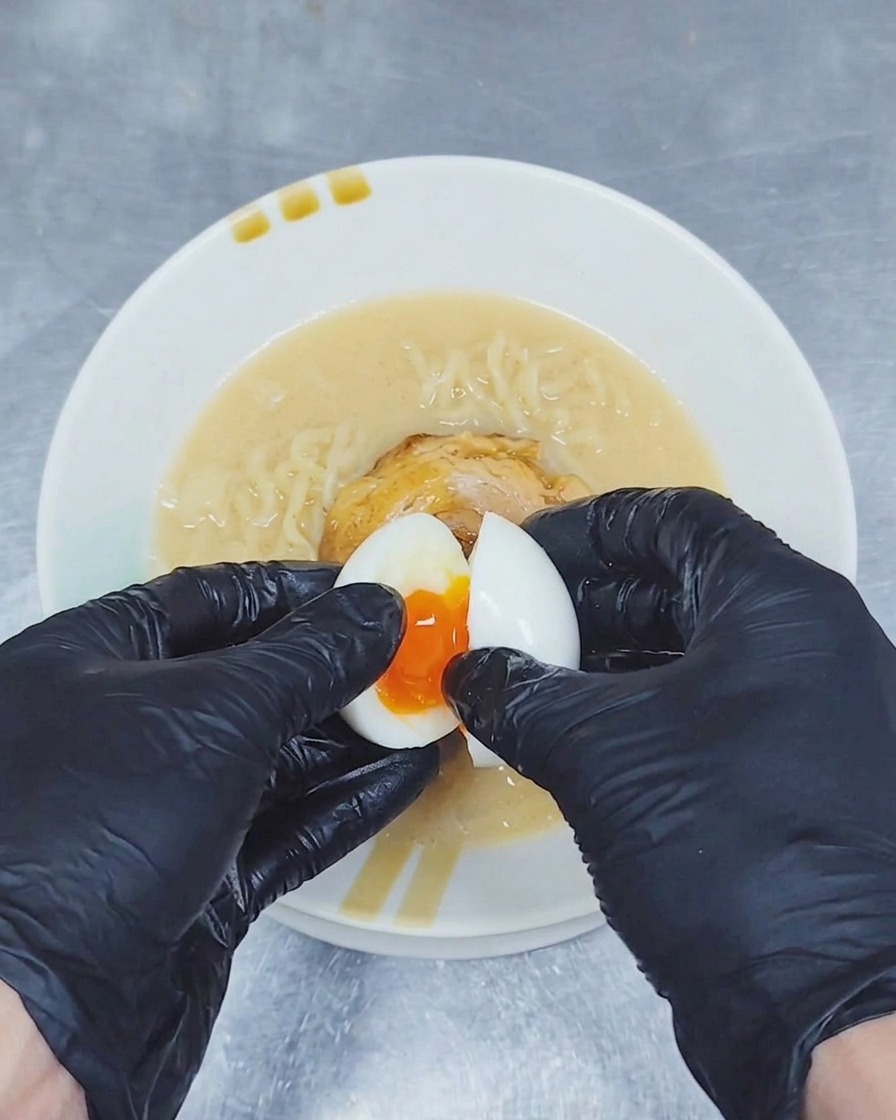 A pair of gloved hands splitting a soft-boiled egg over a bowl of ramen to reveal a runny, golden yolk.