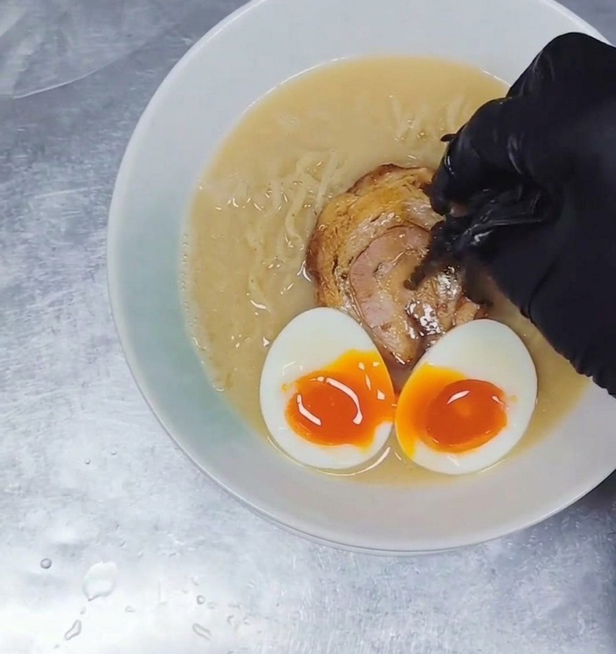 Thin strips of dark wood ear mushrooms being added to a bowl of ramen filled with broth, noodles, pork, and soft-boiled eggs.