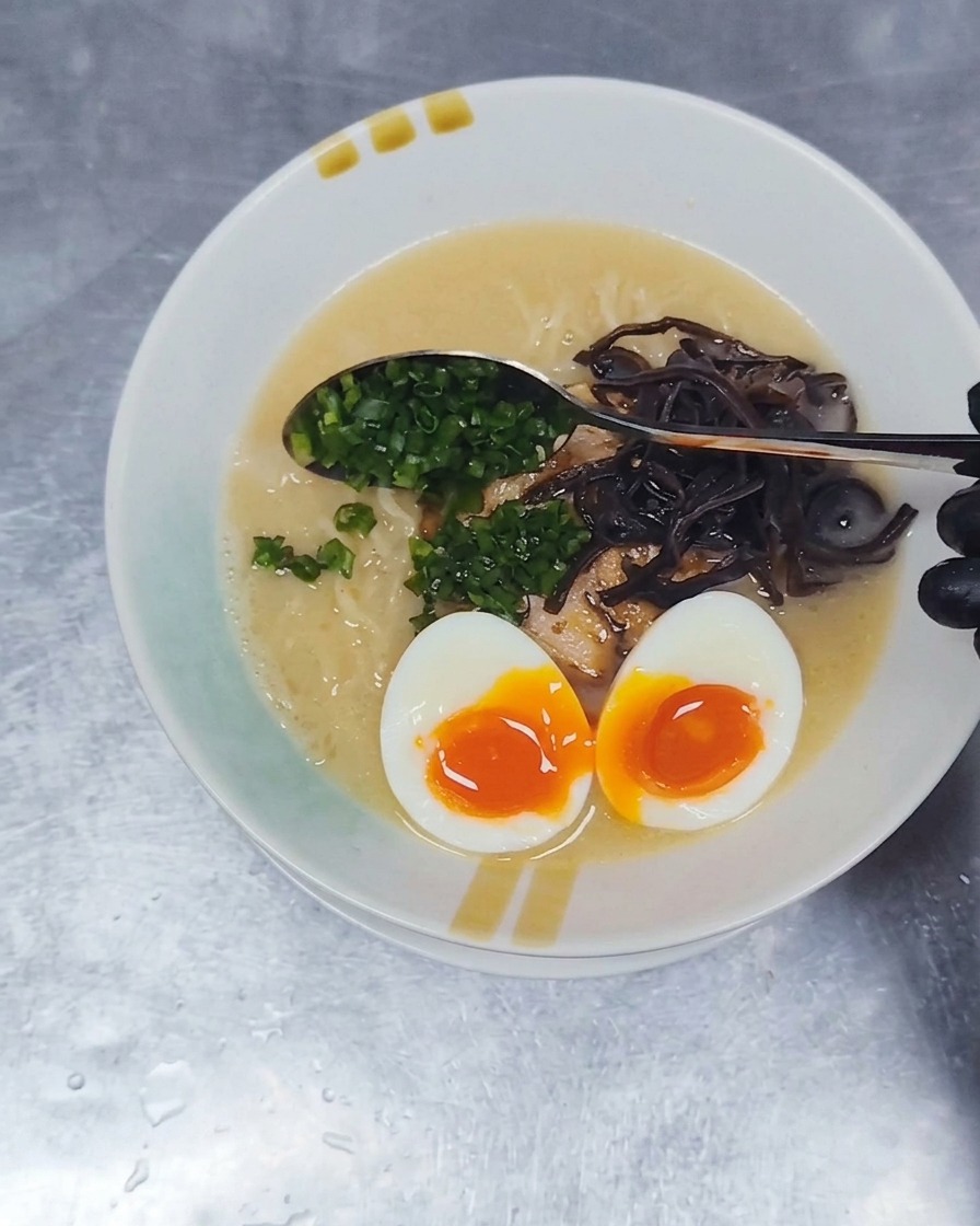 A metal spoon sprinkling finely diced green scallions onto the surface of a Tonkotsu ramen bowl.