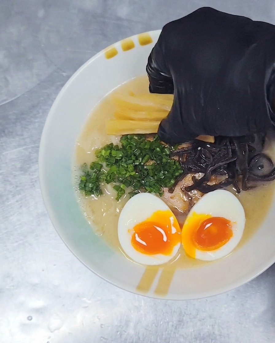 A gloved hand placing yellow strips of seasoned bamboo shoots onto a bowl of ramen already topped with pork, egg, and green onions.