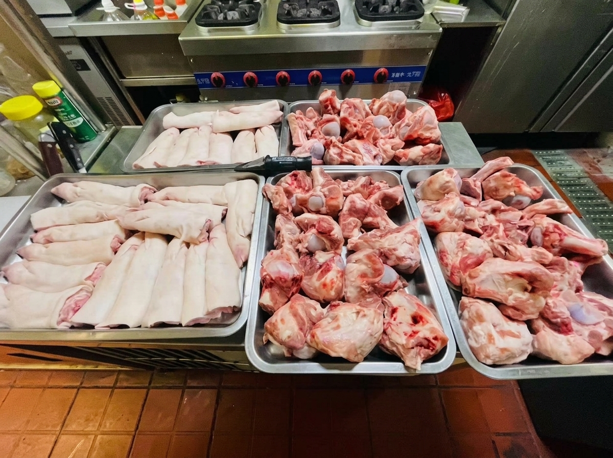 Four metal trays filled with raw pink pork knuckles and trotters arranged on a stainless steel kitchen counter.