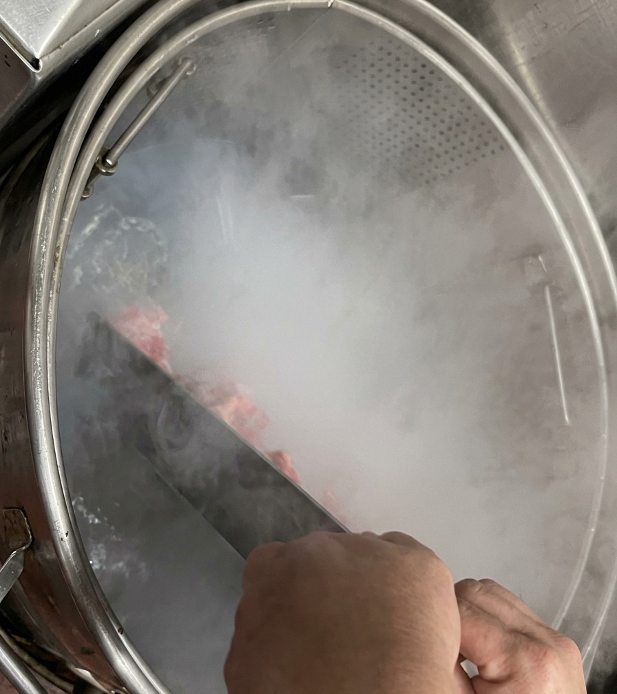Hands holding a large metal stirring paddle, agitating pork bones inside a steaming industrial boiling pot.