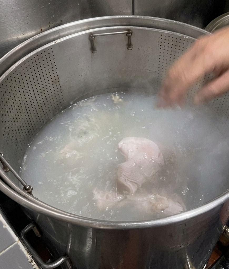 Pork bone pieces submerged in a large pot of boiling water that is turning from clear to a cloudy, milky white.