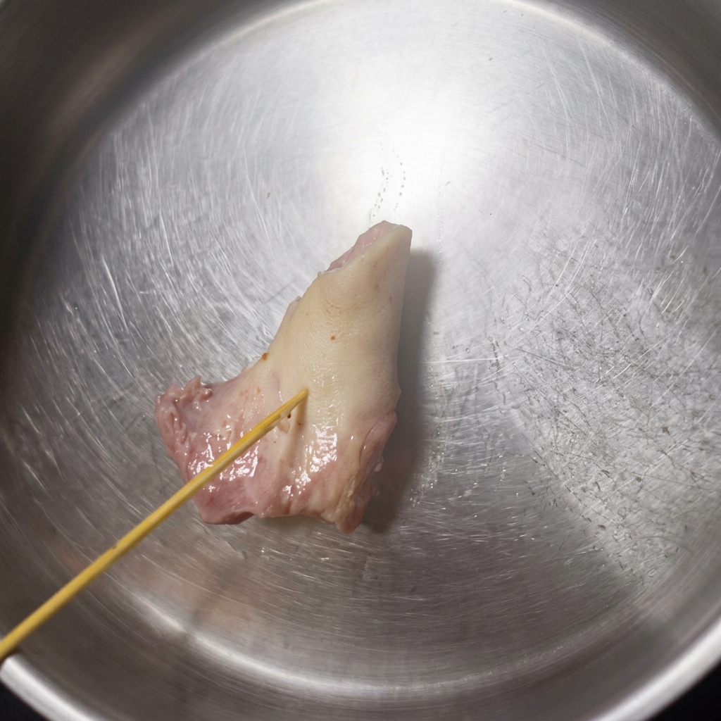 A wooden skewer pressing into a pale, softened pork bone inside a stainless steel bowl, showing the bone beginning to yield.