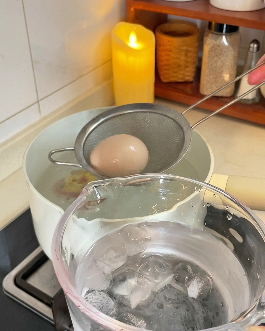 A small metal strainer lifting a brown boiled egg from a white pot and moving it toward a glass cup filled with ice and water.