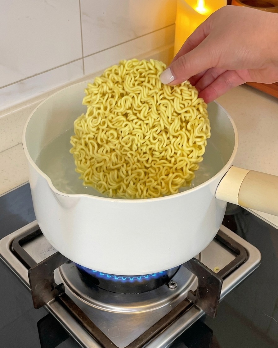 A hand placing a circular block of dry, wavy instant ramen noodles into a white pot of boiling water on a gas stove.
