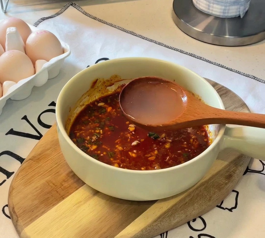 A wooden ladle pouring clear hot water into a light-colored bowl containing a dark reddish-brown seasoning paste.
