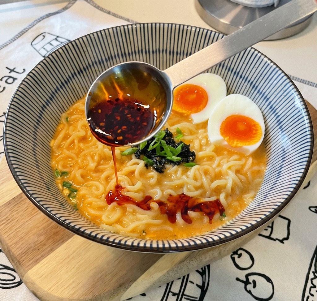 A metal measuring spoon pouring bright red chili oil with visible chili flakes into a bowl of prepared ramen.